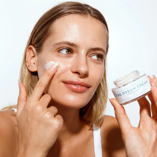Woman holding a jar of The Dream Cream moisturizer from The Organic Skin Co. and applying it to her face 